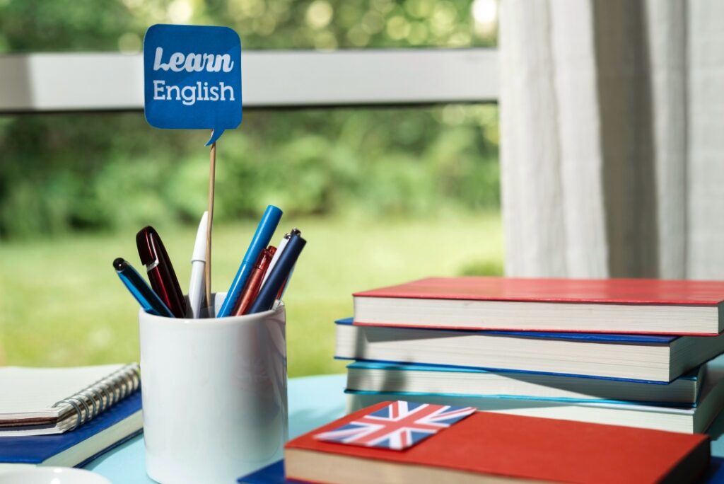 biurko na tle okna a na nim książki oraz kubek z długopisami i flaga z napisem learn english pociąg do angielskich phrasali Preston Publishing