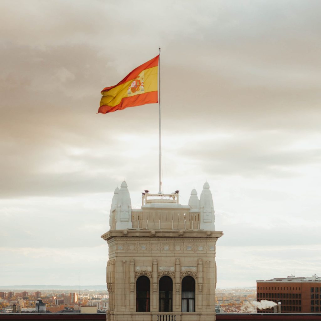 flaga hiszpańska powiewająca na wietrze na szczycie zabytkowej wieży z widokiem na miasto Hiszpańskie tradycje i zwyczaje Preston Publishing
