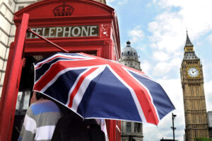 widok na Londyn w tle Big Ben czerwona budka telefoniczna na pierwszym planie ktoś stoi przed budka pod parasolka na ktorej jest wzór flagi brytyjskiej nauka czasów angielskich z książka angielski w tłumaczeniach od Preston Publishing
