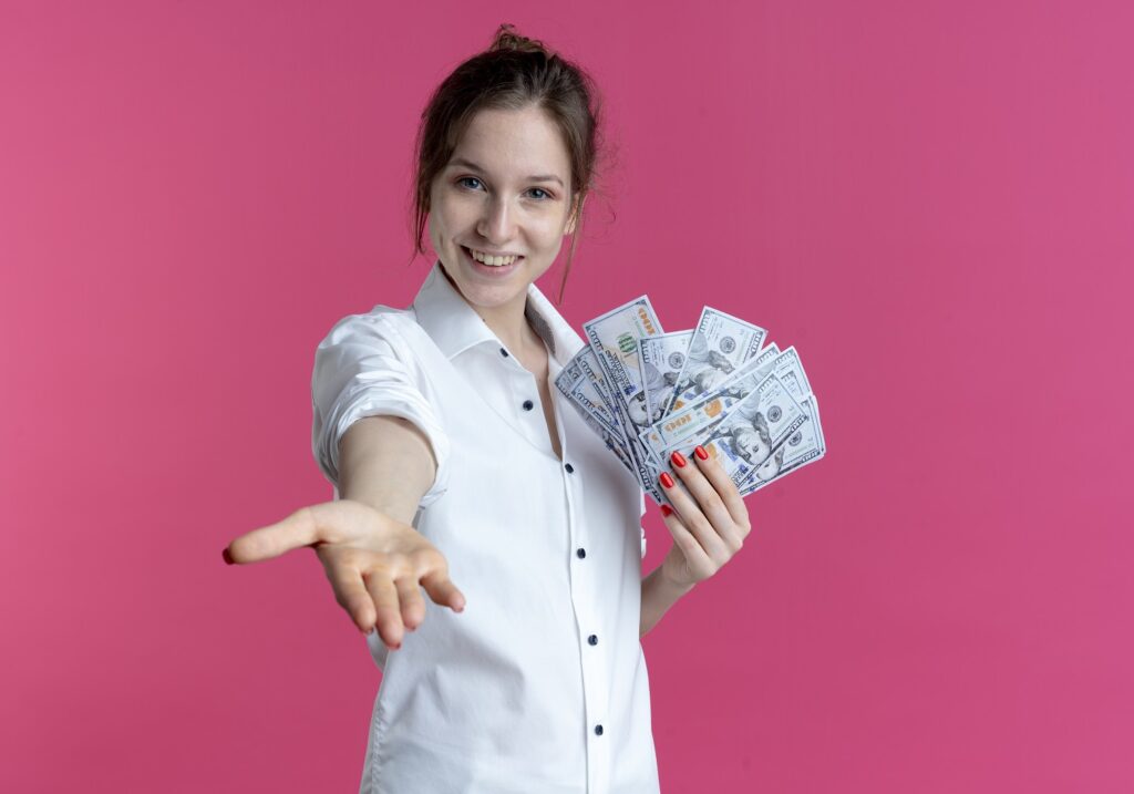 uśmiechnięta młoda kobieta w białej koszuli trzymająca w jednej ręce banknoty jak wachlarz a drugą wyciągnięta do przodu na rozowym tle borrow lend angielskie pulapki jezykowe Preston Publishing
