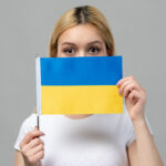 blondynka w białej bluzce na szarym tle trzymająca w rękach mała flagę ukraińska zasłaniająca część twarzy polski_dla_ukraincow_jezyk-polski-w-tlumaczeniach_sytuacje_Preston_Publishing
