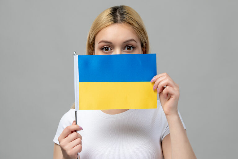 blondynka w białej bluzce na szarym tle trzymająca w rękach mała flagę ukraińska zasłaniająca część twarzy polski_dla_ukraincow_jezyk-polski-w-tlumaczeniach_sytuacje_Preston_Publishing