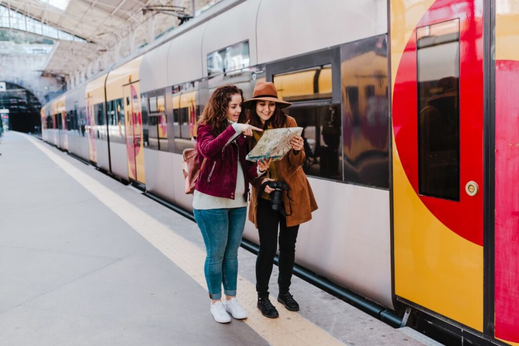 dwie kobiety z plecakami stojące na peronie dworca kolejowego i patrzące na mapę a na peronie stoi pociąg nauka hiszpańskiego zwroty na dworcu i w podróży Preston Publishing