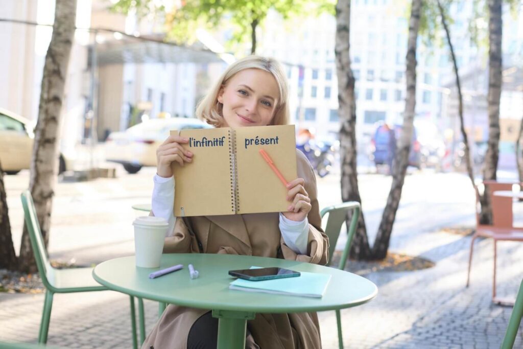 uśmiechnięta dziewczyna w kawiarni francuskiej pokazuje notatki w zeszycie i napis linfinitif prezent bezokolicznik w języku francuskim Preston Publishing