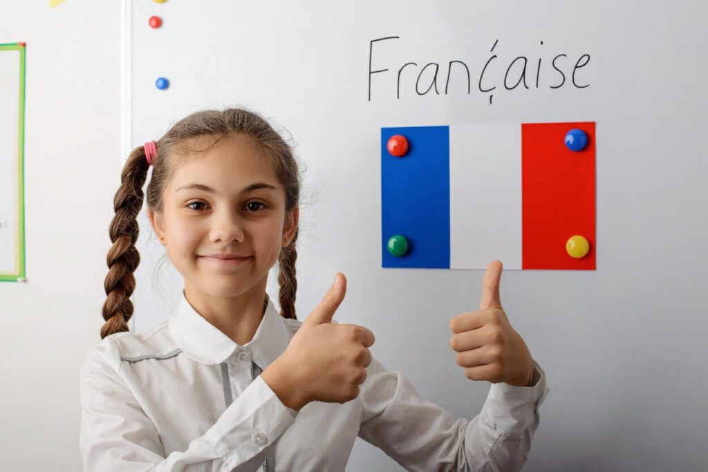 dziewczynka z warkoczykami z kciukami uniesionymi w gore na tle tablicy z napisem francais i flaga francuska nauka języka francuskiego bezokolicznik Preston Publishing