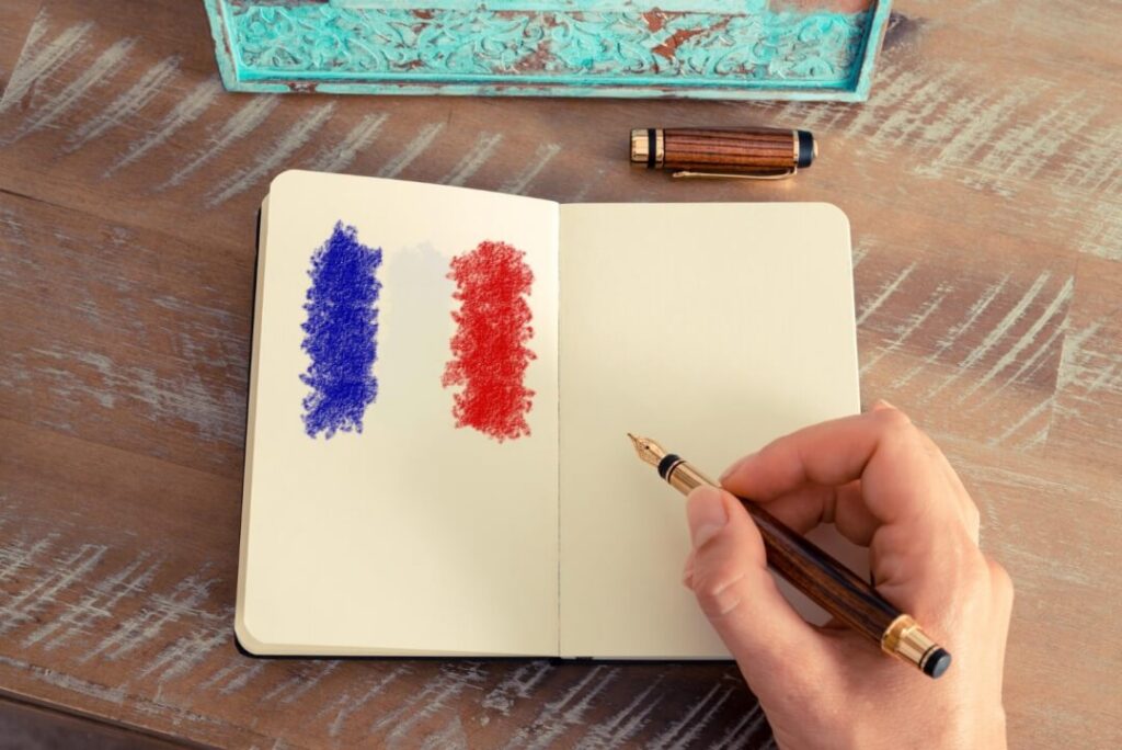 notes z narysowana flaga francuska oraz ręką z długopisem przymierzająca się do pisania imiesłowy w języku francuskim le participe présent Preston Publishing