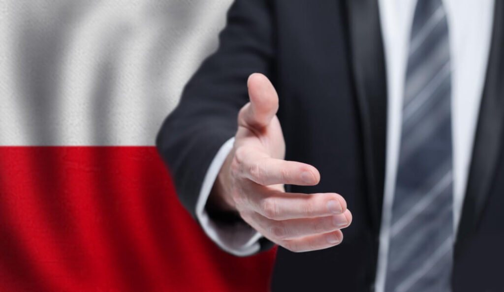 mężczyzna wyciągający dłoń do powitania na tle flagi polskiej nauka polskiego podstawowe zwroty i słówka Preston Publishing