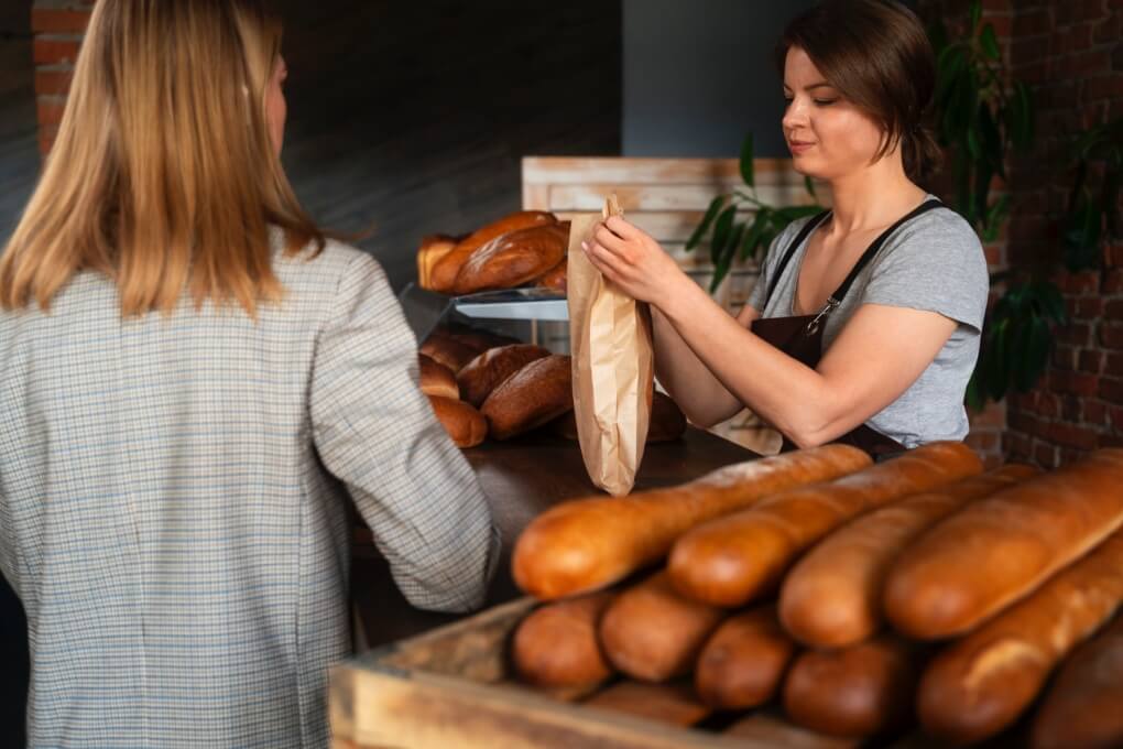 kobieta kupuje bagietkę we francuskiej piekarni a ekspedientka pakuje bagietkę do torby nauka przydatnych zwrotów po francusku przy zakupach Preston Publishing