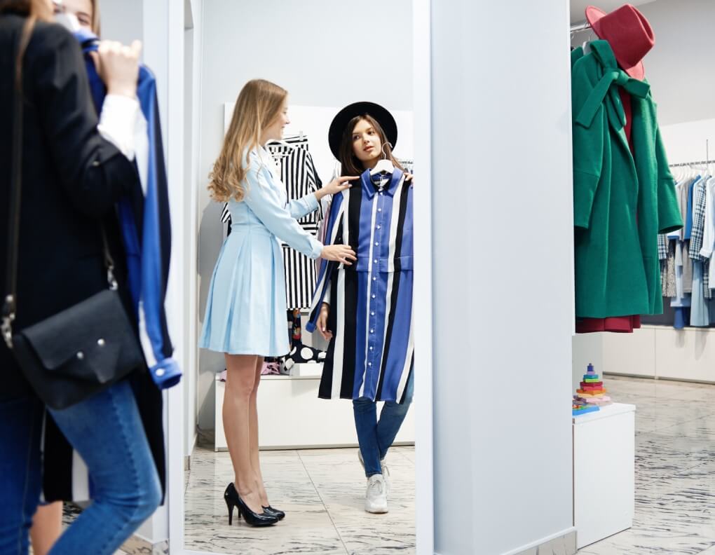 kobieta w sklepie rozmawia z asystentka o rozmiarze ubrania przyjkladajac do siebie sukienkę na wieszaku Preston Publishing