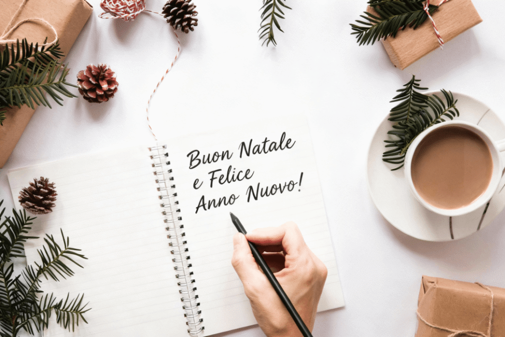 ręką pisząca życzenia świąteczne po włosku w notesie na biurku przyozdobionym świątecznymi szyszkami i gałązkami oraz z kawa w filiżance nauka włoskiego z Preston Publishing