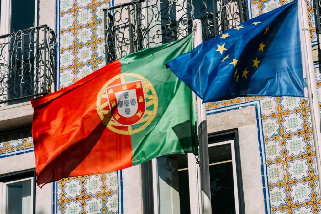 dwie flagi powiewajace na wietrze portugalska i flaga unii europejskiej na tle budynku z portugalskimi plytkami azulejos nauka portugalskiego Preston Publishing